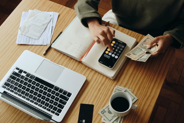 Como-criar-um-planejamento-financeiro-que-funciona-de-verdade-mesade-madeira-com-notebook-dinheiro-com-xicara-de-cafe-em-cima-caderno-com-celular-em-cima-com-pessoa-calculando.jpg