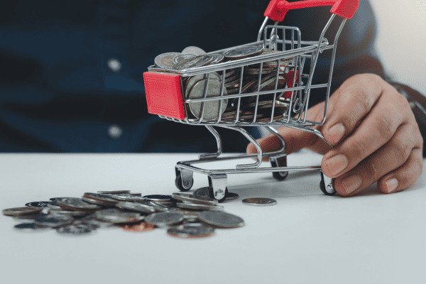 Mão segurando mini carro de compras cheio de moedas e am cima de mesa com mais algumas moedas ao lado